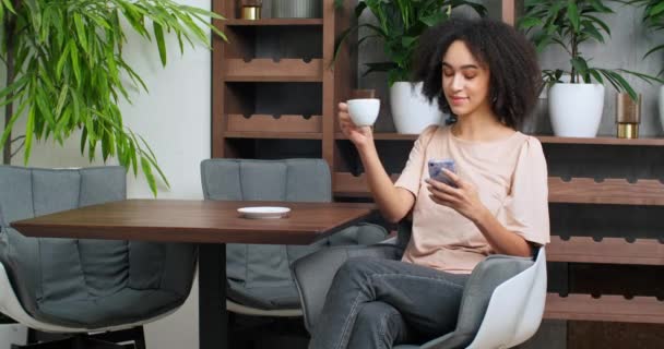 Afro americano étnico rizado mujer adolescente bebiendo café de taza blanca disfrutando de té de capuchino caliente mirando en el teléfono inteligente móvil utiliza dispositivo gadget para la comunicación de chat de navegación en la cafetería — Vídeos de Stock