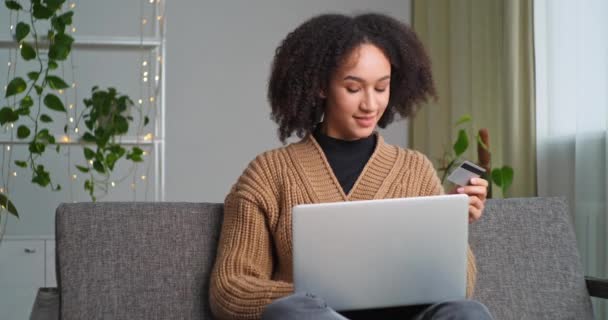 Afroamerikanerin sitzt zu Hause mit Laptop beim Online-Shopping hält Kreditkarte Buchbestellung freut sich Empfangen Geschenkbox von Kuriermann Hände in Latex-Handschuhen, Lieferservice während Sperrung — Stockvideo
