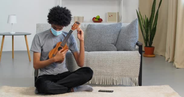 Jovem adolescente afro-americano negro artista músico sentado em casa no chão vestindo máscara médica tocando pequeno ukulele aprendendo a criar melodias músicas ouvindo som, bloquear o conceito — Vídeo de Stock