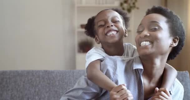 Afroamerikansk ung kvinna mamma äldre syster barnflicka sitter på soffan hemma krama liten söt barn förskola flicka rolig poserar framför kameran tillsammans leende tänder fnissa brett, ansikte närbild — Stockvideo