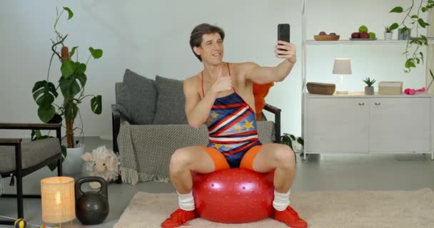 Αστείος καυκάσιος αθλητής σέξυ άνδρας με ρετρό κοστούμι που κάθεται πηδώντας πάνω σε μπάλα χωράει smartphone σε κωμική στάση βάζει τον αντίχειρα επάνω δείχνει δύο δάχτυλα σύμβολο ειρήνης κάνει αυτο φωτογραφία στο τηλέφωνο κάμερα — Αρχείο Βίντεο