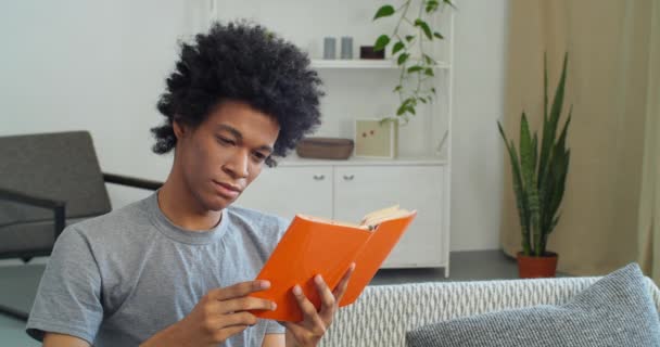Porträtt av afro amerikanska tonåring pojke kille student med lockigt hår sitter i hemmet interiör läsning orange bok vänder sida studie med litteratur lärobok studier information åtnjuter hobby på fritid — Stockvideo