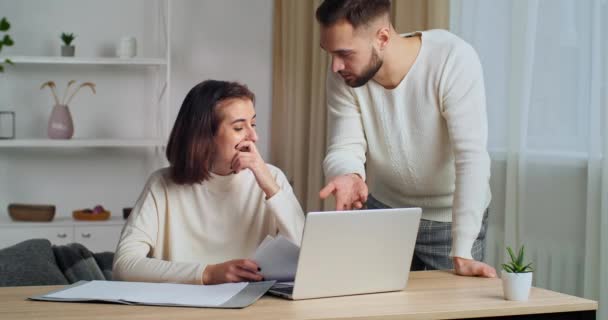 Jonge vrouw vrouw zit aan tafel kijkt naar laptop heeft problemen met papierwerk maakt fout in document serieuze man benaderingen argumenteert verontwaardigd leest brief voelt woede op domme meisje — Stockvideo