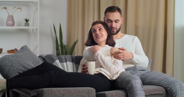 Счастливые кавказская семья пара расслабляющий разговор, смех держа чашки пить кофе чай сидя на диване вместе в гостиной, любящий бородатый муж и жена связи наслаждаясь приятным разговором — стоковое видео