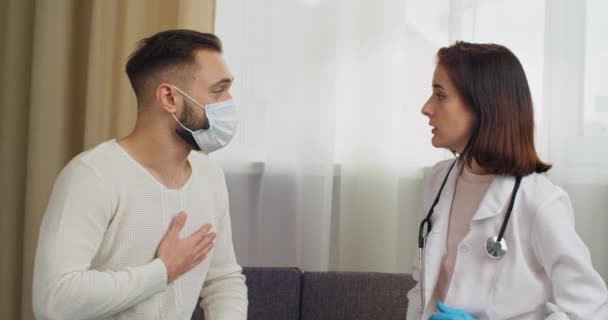 Beteg kaukázusi fiatalember visel orvosi arc maszk konzultál hivatásos női orvos panaszkodik mellkasi fájdalom fejfájás, nő terapeuta méri a hőmérséklet a beteg a tenyerével kesztyűben — Stock videók