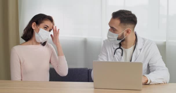 Άνδρας γιατρός με ιατρική στολή κάθεται στο τραπέζι στο νοσοκομείο κλινική λαμβάνει συμβουλές γυναίκα ασθενής γυναίκα σε ιατρική μάσκα ακούει τα συμπτώματα των καταγγελιών δακτυλογράφηση για τη διάγνωση laptop συνταγογραφεί θεραπεία — Αρχείο Βίντεο