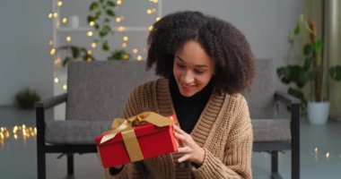 Güzel, mutlu, meraklı, Afro-Amerikalı bir kadın, evinde, rahat oturma odasında oturmuş elinde altın kurdeleyle kırmızı bir hediye kutusu tutuyor. Hediyeler, içkiler, tatil konsepti.