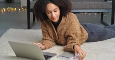 Ciddi ciddi sorumlu etnik kadın dizüstü bilgisayara bakıyor, yerde yatan Afro-Amerikan kız öğrenci bilgisayarını kullanarak evden internet yazma sınavına çalışıyor.