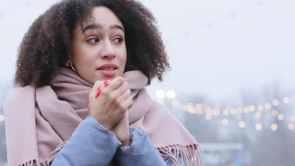Portret afro amerykańskiej kobiety z kręconymi włosami nosi ciepłe ubrania kurtka i szalik stoi sam w zimną pogodę w mieście ogrzewa jej ręce masuje dłonie dmuchając gorące powietrze na palce z doskonałym manicure — Wideo stockowe
