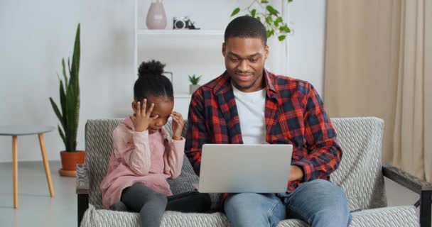 Afro amerikansk far med dotter äldre bror med lillasyster sitter på soffan titta på laptop skräckfilm, barn blundar med händerna i rädsla och ser eftertänksamt på datorskärmen — Stockvideo