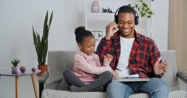 Lustige fürsorgliche afroamerikanische Tochter kleines Mädchen sitzt mit Papa auf Sofa schließt Laptop des geschäftigen Geschäftsmannes trägt Kopfhörer Pausen bei der Arbeit Praktiken Meditation Lotusposition lindert Stress Ruhe — Stockvideo