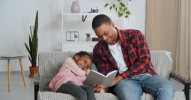 Genç baba sevgili kızına kitap okuyor. Fotoğraflar gösteriyor. Çocuk oturma odasında oturuyor, Afrikalı Amerikalı erkek ebeveyn küçük kıza okumayı öğretiyor, aile evde gülüyor.
