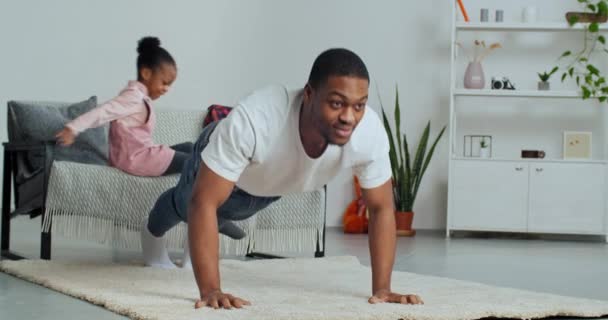 Afroamerikanische Alleinerziehende, die zu Hause Spaß haben, starker Vater, der Sport treibt, während er sich auf dem Teppich vom Boden nach oben schiebt, während unerwartet geliebte Tochter ihrem Papa auf den Rücken springt und ihn umarmt — Stockvideo