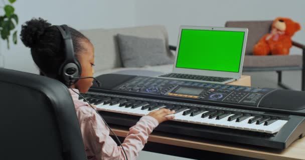 Afro fille enfant jouant sur synthétiseur porte des écouteurs regardant dans un ordinateur portable avec écran vert écoute leçon de pratique de la musique. Vue de derrière enfant crée une chanson en utilisant des touches de piano électronique — Video