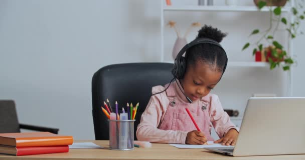 Concentré afro américaine petite fille enfant assis à la maison à la table dessine l'image choisit crayon porte casque écoute professeur Internet regarder la vidéo leçon sur ordinateur portable chat conférence en ligne — Video