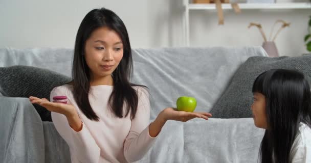亚洲单身母亲坐在家里，女儿手里拿着饼干和苹果，双手拿着平衡的姿势向孩子们表示选择的女孩拒绝烘焙糖果，吃着健康的水果，养成了良好的习惯 — 图库视频影像