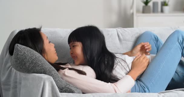 Щасливий безтурботний азіатська корейська мати і маленька дитина дочка лежить на дивані диван сміється весело посміхаючись разом, дитина цілує маму на носі, насолоджуючись зв'язуванням обіймів вдома у вихідні — стокове відео