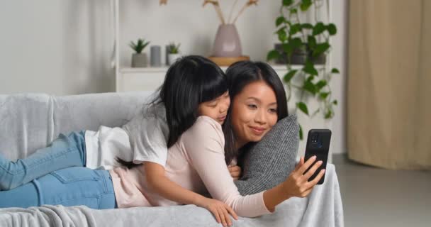 Μικρό ασιατικό κορίτσι αγκαλιάζει τη μητέρα πίσω από την πλάτη της, μαμά κρατά το τηλέφωνο στο χέρι της παίρνει selfie φωτογραφία στο web κάμερα του κινητού smartphone χρήση online app με μάσκες chat στα κοινωνικά δίκτυα βλέποντας βίντεο — Αρχείο Βίντεο