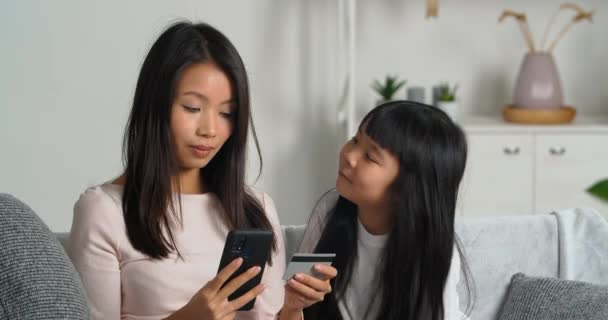 Asyalı bir ailenin genç annesi ve sevgili kızı modern oturma odasında birlikte oturuyorlar. Annesi elinde telefon ve kredi kartı tutuyor ve evine çevrimiçi alışveriş siparişleri gönderiyor. — Stok video