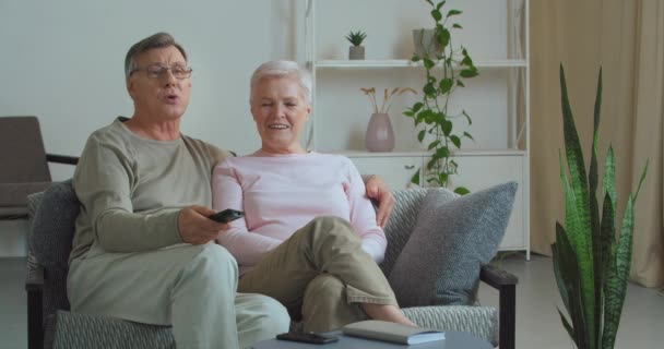 Пожилая кавказская пара зрелая семья седовласый мужчина и женщина бабушки и дедушки сидят на диване интерьер гостиной обнимаясь смотреть телевизор камера муж переключения каналов с помощью пульта дистанционного управления — стоковое видео