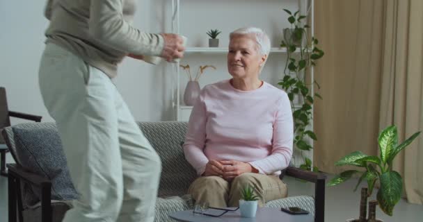 Viejo trae a su amada esposa taza de té caliente café capuchino cuidar de cónyuge femenino. cariñosa madura abuelita pareja relajarse en acogedor sofá por la mañana. Amante hombre de mediana edad con mujer de edad avanzada — Vídeo de stock