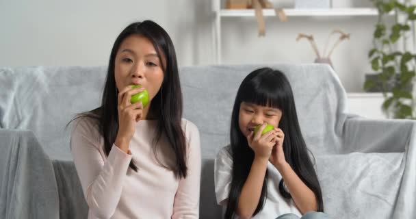 Ung asiatisk familj två personer mor och dotter flicka sitter hemma vardagsrum äta gröna saftiga sura äpplen njuta säsongsbetonad frukt äta vitamin bantning torka varandra kinder — Stockvideo