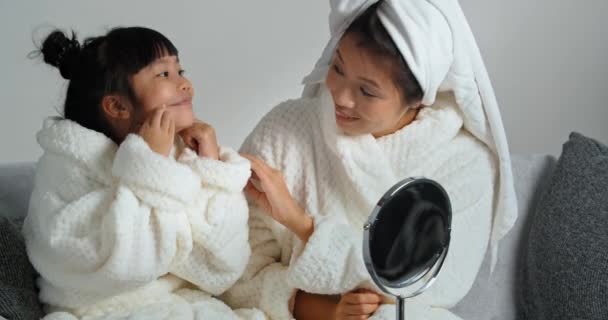 Азійська дівчинка в халаті і корейській етнічній жінці, яка сидить разом на кушетці після вечірньої спа-дощу дивлячись на відображення в дзеркалі дитини гримати, в той час як мама випрямляє волосся. — стокове відео