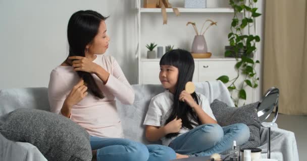 Молода азіатська жінка самотня мати вчить кохану маленьку доньку дівчинку розчісувати довге темне волосся показує, як використовувати пензлик, щоб доглядати за за зачіскою вдома, стильна концепція, приготування ранкової краси — стокове відео