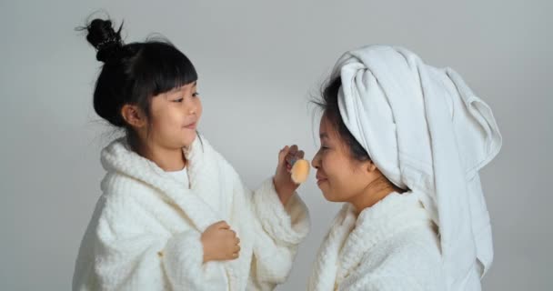 Χαριτωμένο μικρό ασιατικό κορίτσι κόρη και η μαμά φορούν μπουρνούζια μετά το μπάνιο spa απολαμβάνουν καθαριότητα και φρεσκάδα. Παιδί κάνει μακιγιάζ για τη μητέρα χρησιμοποιώντας καλλυντικά φιλιά βούρτσα γυναίκα στη μύτη, αγάπη έννοια — Αρχείο Βίντεο