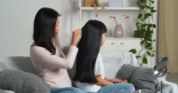 Щаслива любляча азійська сім'я. Корейська мати носить повсякденний одяг, який поєднує її дочок з волоссям, використовує гребінь, щоб піклуватися про її доньку-брюнетку, яка сидить на дивані у вітальні. Любов до сім "ї — стокове відео