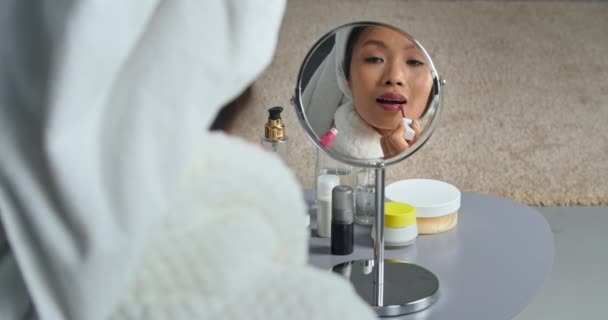 Αντανάκλαση νεαρή όμορφη ασιατική κορεάτικη γυναίκα εθνοτικής κορίτσι κοιτάζοντας στον καθρέφτη φοράει λευκό μπουρνούζι και πετσέτα στο κεφάλι της βάφει τα χείλη της με κραγιόν κάνει μακιγιάζ ετοιμάζεται για εκδήλωση, κοσμετολογία — Αρχείο Βίντεο