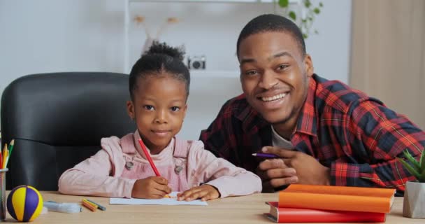Ung far lärare afro amerikansk kille med söt älskad dotter skolflicka sitter vid bordet ritning, pappa hjälper liten flicka med bild kontroller läxa tittar på kameran viftar med händerna i hälsning — Stockvideo