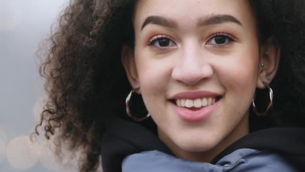 Vista frontal de chica afroamericana con maquillaje natural lleva pendientes de joyería se encuentra en el fondo de la ciudad de invierno y caminar a la gente mirando a la cámara sonriente, primer plano de la cara femenina con piel oscura — Vídeo de stock