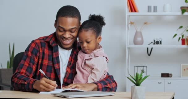 Афро-американський неодружений батько перевіряє домашнє завдання, допомагаючи милій школярці з навчанням вчити дитину писати, сидячи за столом у вітальні. Приватна освіта під час блокування — стокове відео