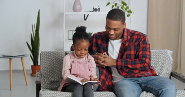 Afro amerikanska pappa sitter i vardagsrummet med sin dotter lyssnar på agirl barn läser bok saga berättelse att studera hemma under lockdown och karantän, unga far hjälper barnet med att lära — Stockvideo