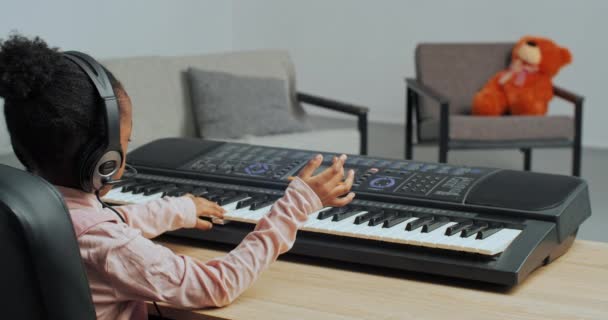 Enfant afro-américain méconnaissable petite fille porte des écouteurs apprenant à jouer du piano à l'aide d'un synthétiseur électrique clavier à la maison, musicien débutant étudiant compose mélodie, vue de derrière — Video
