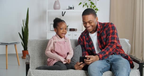 Afro americano hombre padre y pequeña hija sentado en cómodo sofá en acogedor habitación papá muestra bebé video película de dibujos animados en el teléfono hace selfie con divertido máscaras chica cubre su nariz congelada con su mano — Vídeo de stock