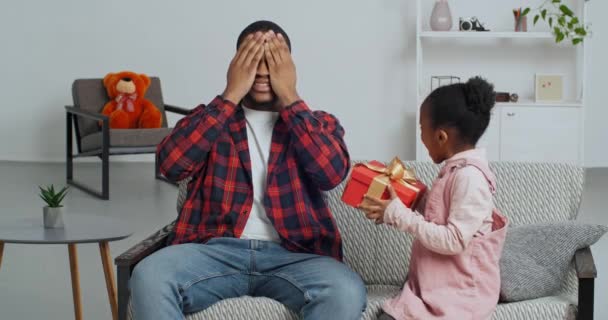 Nettes Faro-Kind Tochter machen Überraschungsgeschenk für Papa erhalten Geschenkbox auf Sofa sitzen, schwarzer Mann Vater bedeckt Augen schließen, während kleines Kind Mädchen gratuliert Eltern am Vatertag Geburtstag zu Hause — Stockvideo