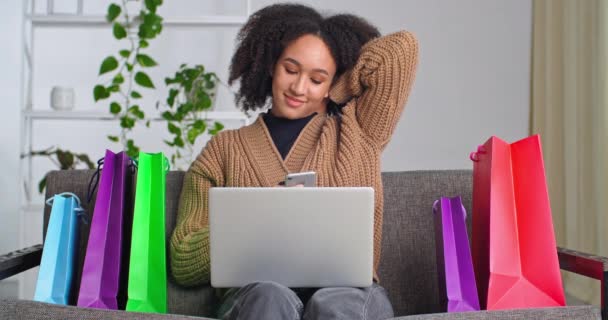 Calma contente menina afro-americana senta-se no sofá na sala de estar ao lado de sacos de presente brilhante compras com laptop olha para celular smartphone bate-papo on-line compra bens on-line faz a ordem olhando para a câmera — Vídeo de Stock