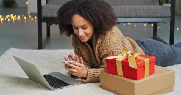 Curly-haired teenage girl freelancer business woman lie at home on floor on white carpet holding phone in her hands looks into laptop uses modern gadgets to order online to home gifts during lockdown — Stock Video