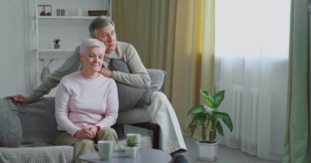 Пожилая пара кавказский старик и седовласая зрелая женщина сидят обнимаясь на диване в современном интерьере гостиной смотреть телевизор говорить наслаждаясь отдыхом вместе в уютном доме — стоковое видео