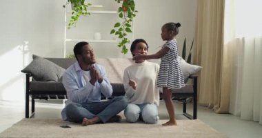 Afro-amerikalı kız anaokulu kızı anne-babası için şarkı söylüyor. Çene hareketleri gösteriyor. Etnik anne ve baba, modern oturma odasında dans eden bir çocuğu dinliyor.
