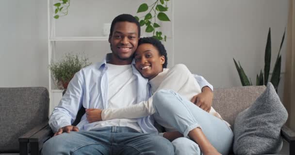 Afro amerikanska par bär casual kläder sitter tillsammans hemma vardagsrum interiör glada nygifta kramar på soffan ler tittar på kameran tillbringa tid tillsammans, helg koppla av, kärlek koncept — Stockvideo