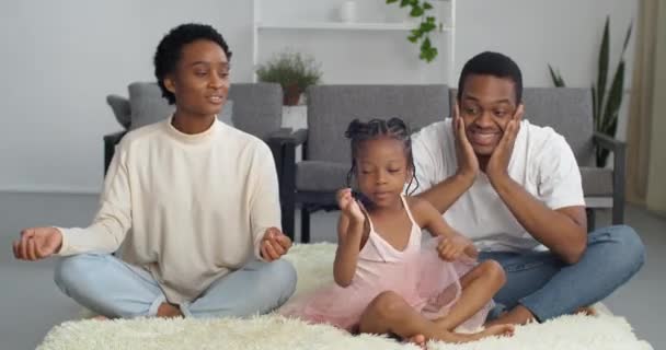 Молодые афро-американские родители учат свою маленькую шумную дочь медитировать, три человека семья сидит в позе лотоса на полу в гостиной чувствовать себя спокойно расслабиться от стресса делать дыхательные упражнения — стоковое видео