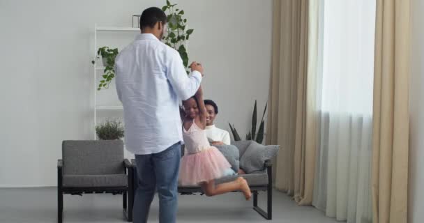 Młody kochający ojciec Afro Amerykanin trzyma za ręce swoją małą uroczą córkę podnosi dziecko do kręgów powietrznych obraca się wokół zabawy z dzieckiem w różowej sukience, matka siedzi na kanapie patrząc na jej rodzinę — Wideo stockowe