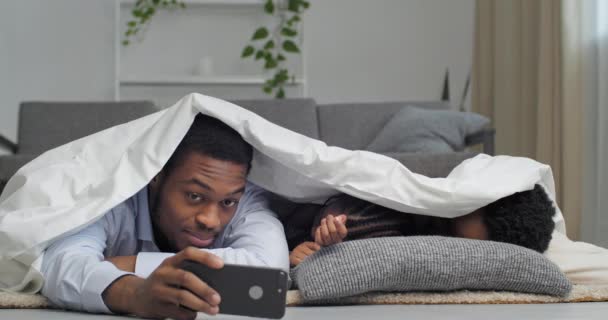 Retrato familiar de los padres afroamericanos con la pequeña hija dormida se encuentran bajo una manta blanca en casa en un acogedor apartamento nuevo, papá toma fotos o videos en la cámara del teléfono móvil con la niña y la esposa — Vídeos de Stock