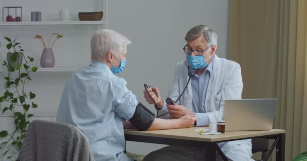 Médico senior en mascarilla facial que mide la presión arterial alta baja de una paciente anciana hospitalizada. Cardiólogo examinando señora jubilada hipertensa usando tonómetro médico en el hospital — Vídeo de stock