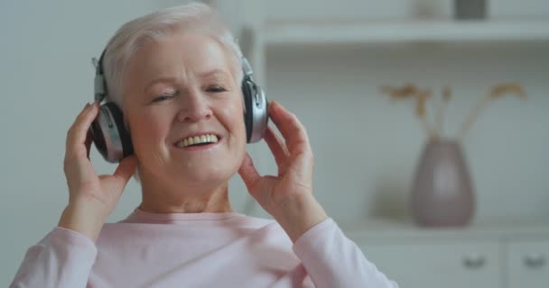 Elderly mature Caucasian lady with gray hair with short hair listening to her favorite music enjoying song using modern headphones moves her head to tune rhythmically having fun at home in retirement — Stock Video