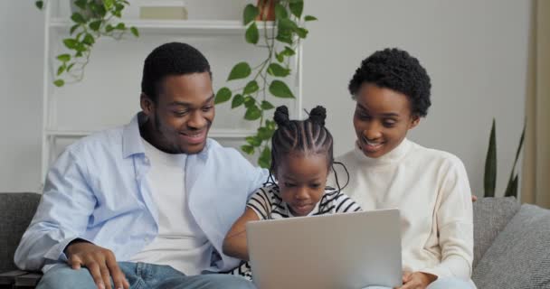 Tres personas padres con hija pequeña pareja afroamericana y linda niña preescolar sentado en el sofá en casa utilizando el ordenador portátil para la comunicación remota video chat conferencias en línea saludos de las manos — Vídeo de stock