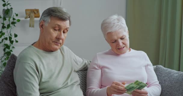 Kaukaskie para stary rodzina siedzi na kanapie w domu salon starsze żona siwe włosy kobieta licząc rachunki dolarów pieniądze dojrzały mężczyzna pokazując karty kredytowe doradza za pomocą elektronicznych finansów, e-biznes — Wideo stockowe